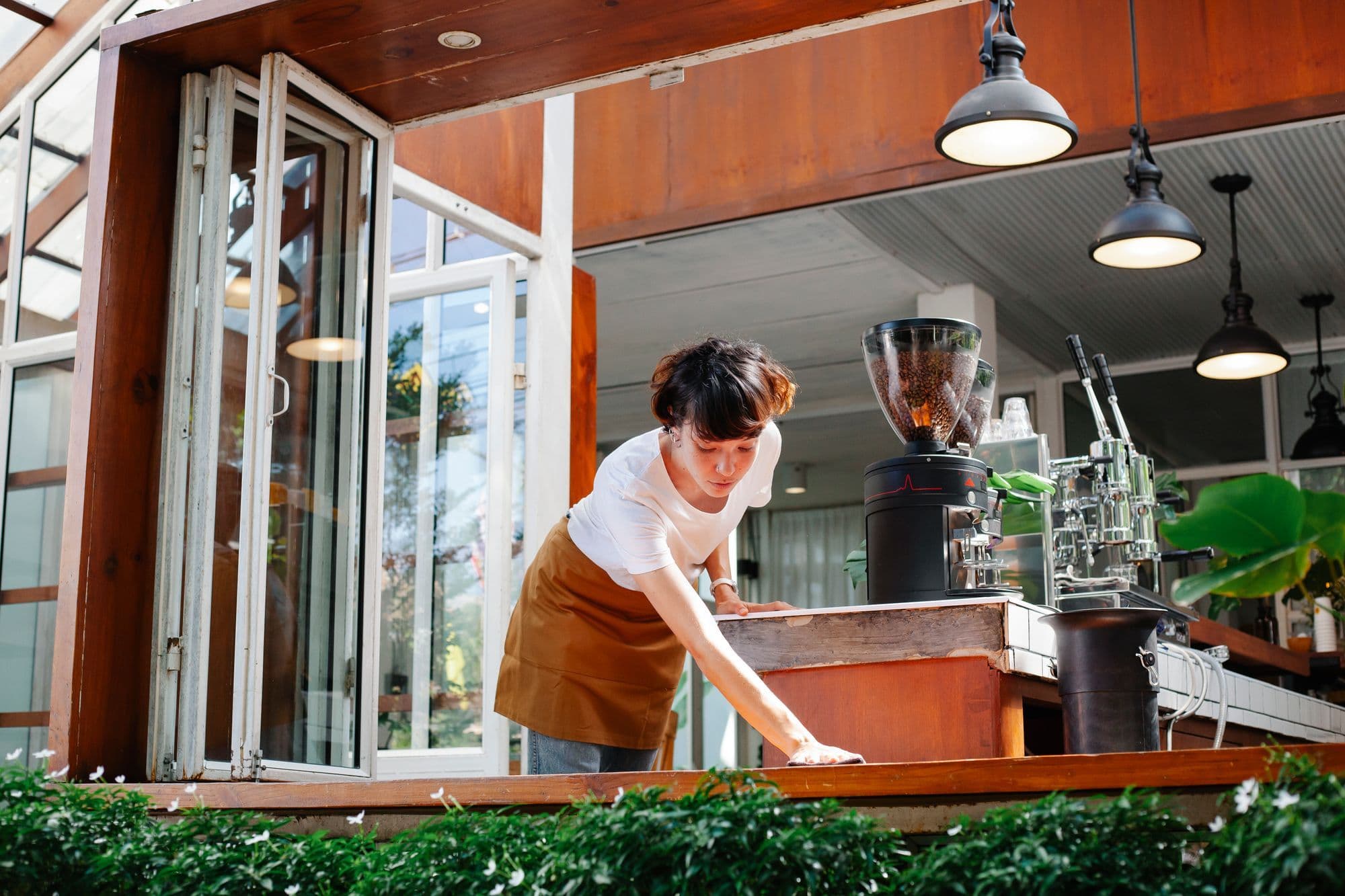 An Asian cafe owner in an apron wiping down the counter of a bright open-window cafe, with an espresso machine and grinder beside her, illustrating the day-to-day reality of opening and running a cafe in Australia.
