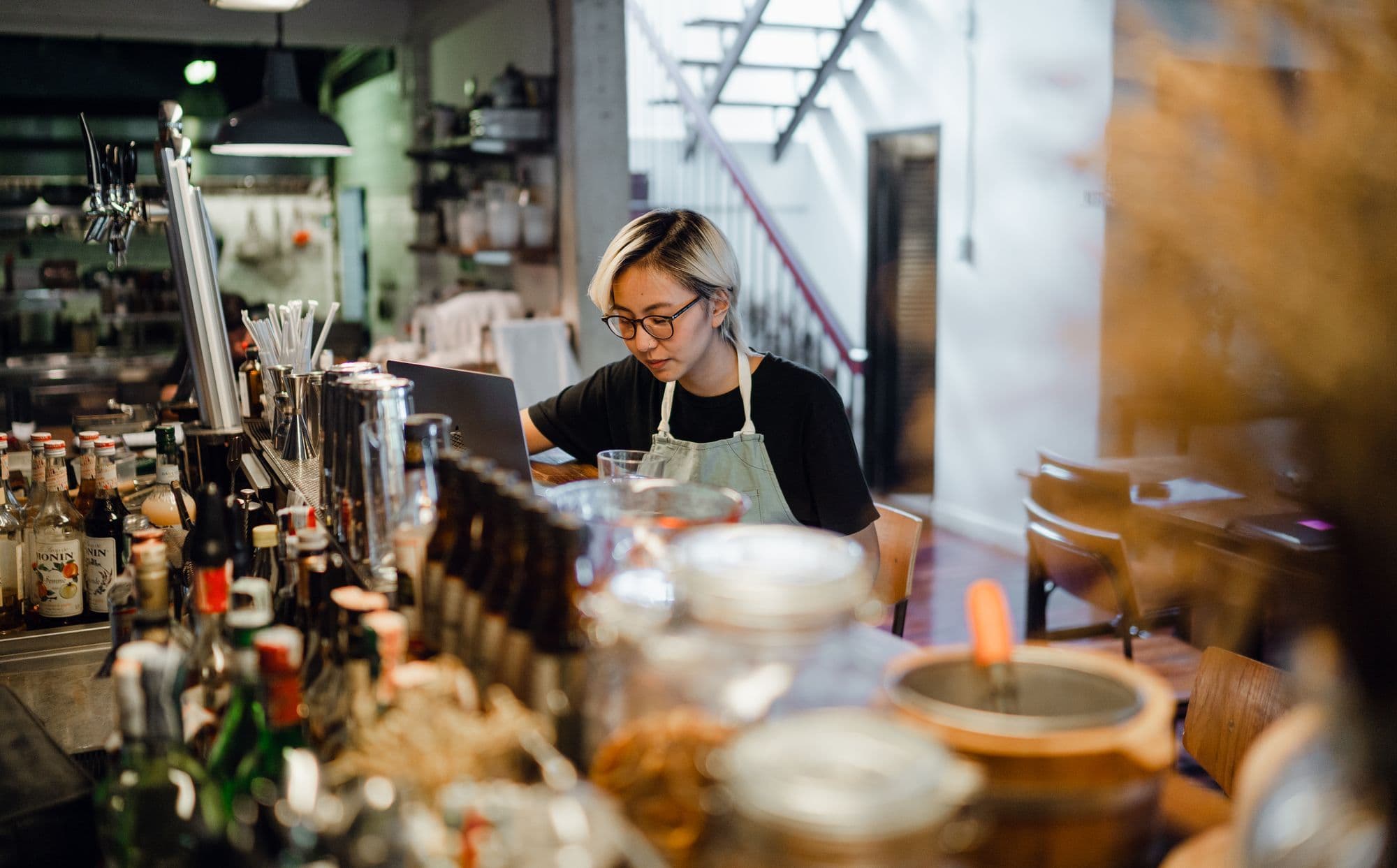 Asian cafe owner in an apron checking their restaurant website on a laptop at the bar counter